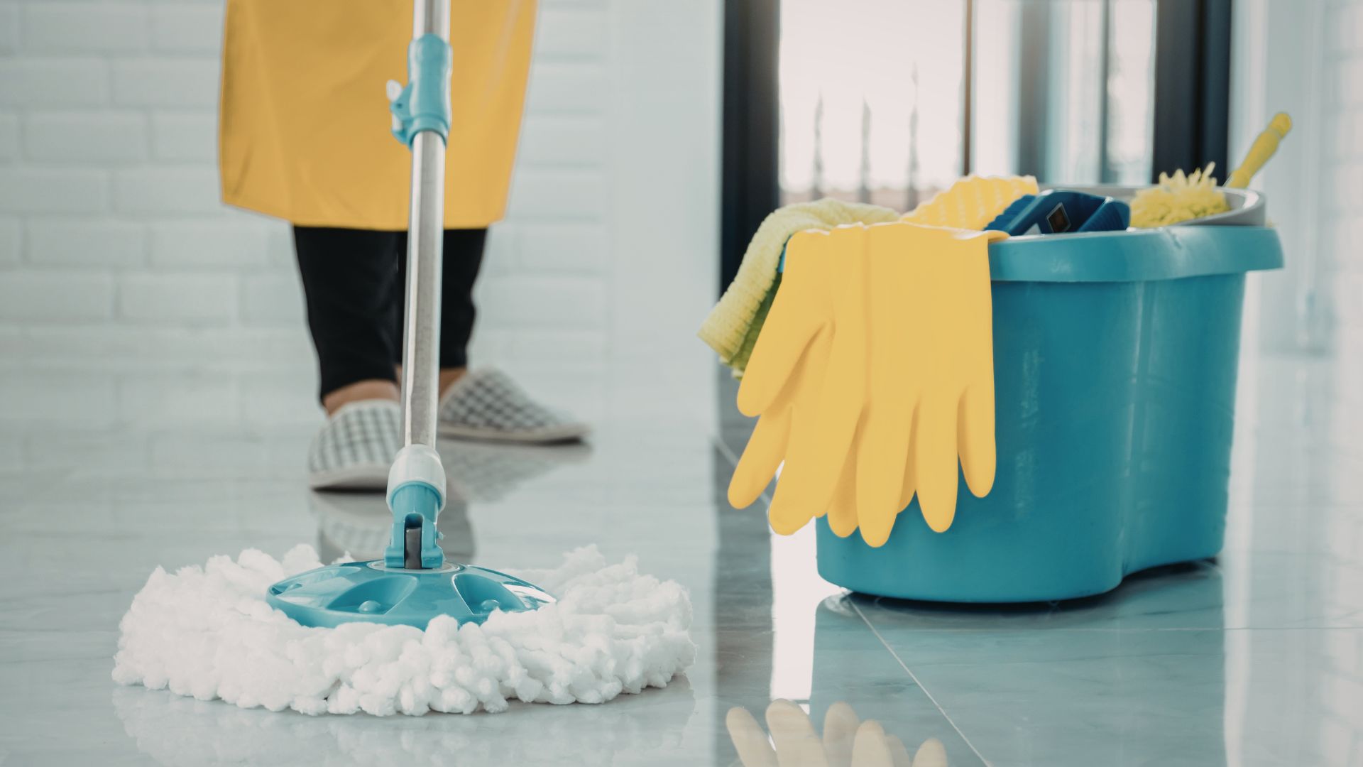 Person in yellow apron and gloves cleaning floor with mop and bucket.